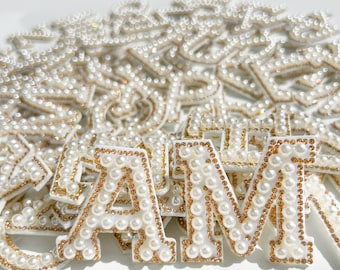 May include: A close-up of a pile of white pearl-embellished letters, including the letters A, M, and a few others. The letters are made of white fabric and have a gold outline.