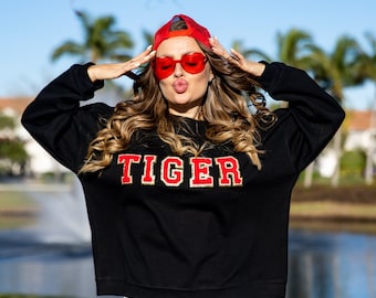 May include: A black sweatshirt with the word "TIGER" in red and gold lettering. The model is wearing a red baseball cap and red heart-shaped sunglasses.