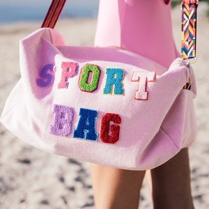 Puede incluir: Una bolsa de deporte rosa con las palabras "SPORT BAG" en letras bordadas de colores.