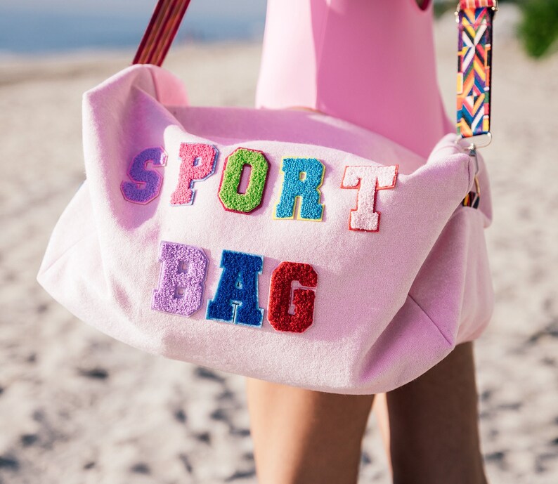 Puede incluir: Una bolsa de deporte rosa con las palabras "SPORT BAG" en letras bordadas de colores.