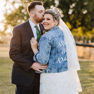 K&ouml;nnte beinhalten: Eine Braut und ein Br&auml;utigam posieren f&uuml;r ein Hochzeitsfoto. Die Braut tr&auml;gt ein wei&szlig;es Hochzeitskleid mit einer Jeansjacke, auf der "Almost There Bride" steht. Der Br&auml;utigam tr&auml;gt einen dunklen Anzug mit einer gr&uuml;nen Krawatte.