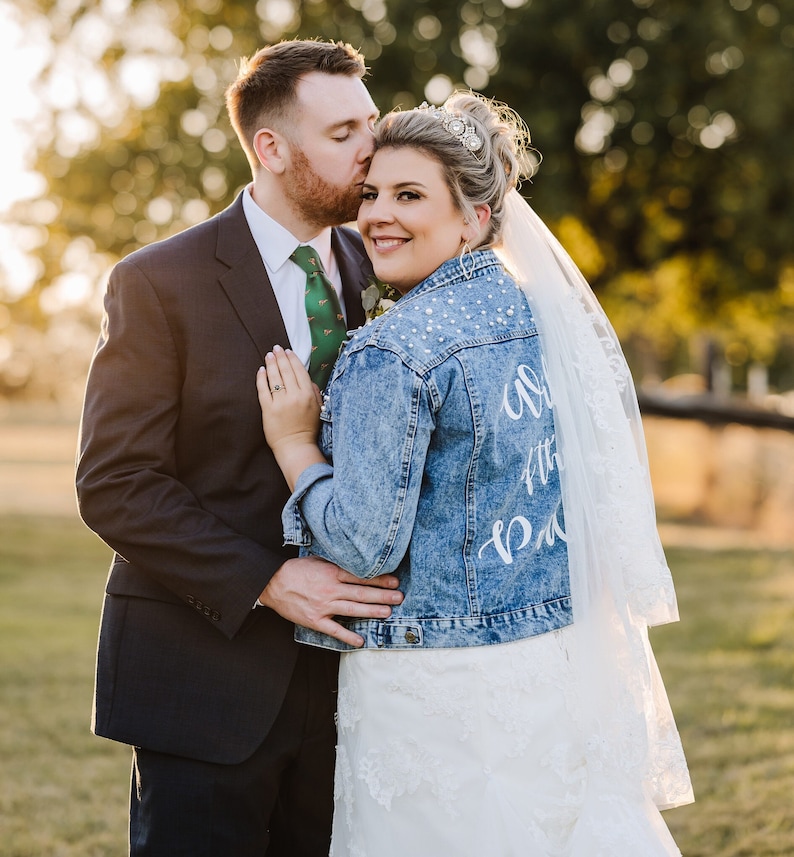 K&ouml;nnte beinhalten: Eine Braut und ein Br&auml;utigam posieren f&uuml;r ein Hochzeitsfoto. Die Braut tr&auml;gt ein wei&szlig;es Hochzeitskleid mit einer Jeansjacke, auf der "Almost There Bride" steht. Der Br&auml;utigam tr&auml;gt einen dunklen Anzug mit einer gr&uuml;nen Krawatte.