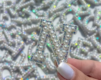 May include: A white pearl letter N, with iridescent accents, is held in a hand. The letter is surrounded by other pearl letters.