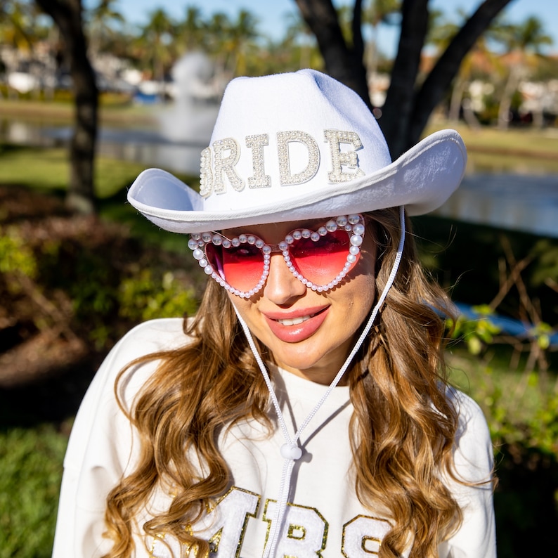 May include: A white cowboy hat with the word "BRIDE" spelled out in rhinestones. The hat is worn by a person wearing pink heart-shaped sunglasses with pearl accents.