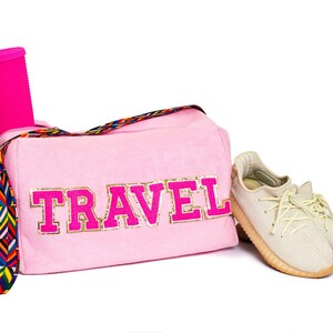 Puede incluir: Bolsa de viaje rosa con una inscripci&oacute;n "TRAVEL" de purpurina dorada y una correa con estampado colorido. Un par de zapatillas beige se encuentra al lado de la bolsa.