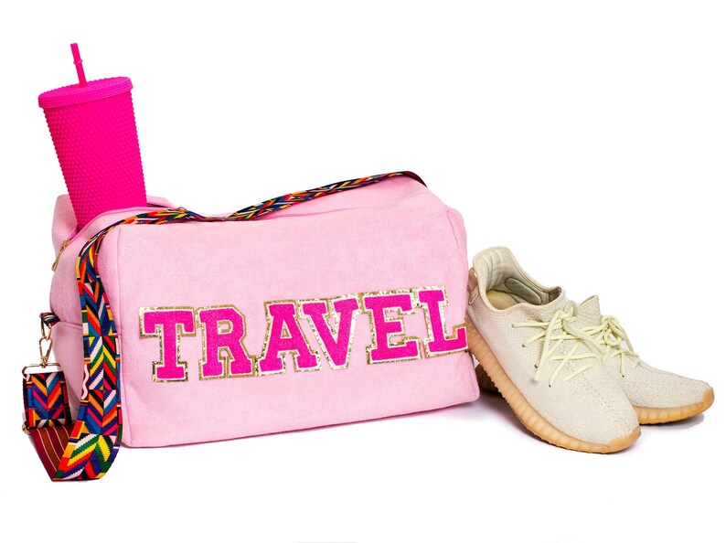 Puede incluir: Bolsa de viaje rosa con una inscripci&oacute;n "TRAVEL" de purpurina dorada y una correa con estampado colorido. Un par de zapatillas beige se encuentra al lado de la bolsa.