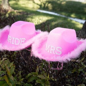 May include: Two pink cowboy hats with rhinestone lettering. One hat says "RIDE" and the other says "MRS". Both hats have a fluffy pink feather trim around the brim. The hats are sitting on top of a bush.
