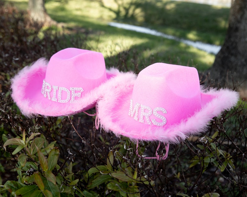 May include: Two pink cowboy hats with rhinestone lettering. One hat says "RIDE" and the other says "MRS". Both hats have a fluffy pink feather trim around the brim. The hats are sitting on top of a bush.