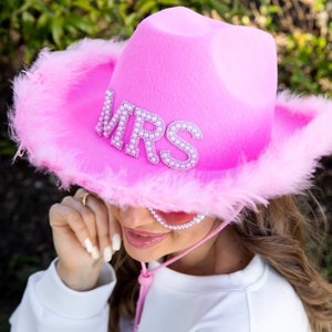 May include: A bright pink cowboy hat with the word "MRS" spelled out in silver rhinestones. The hat has a fluffy, feathered trim and a pink chin strap. A fun accessory for a bachelorette party or wedding.