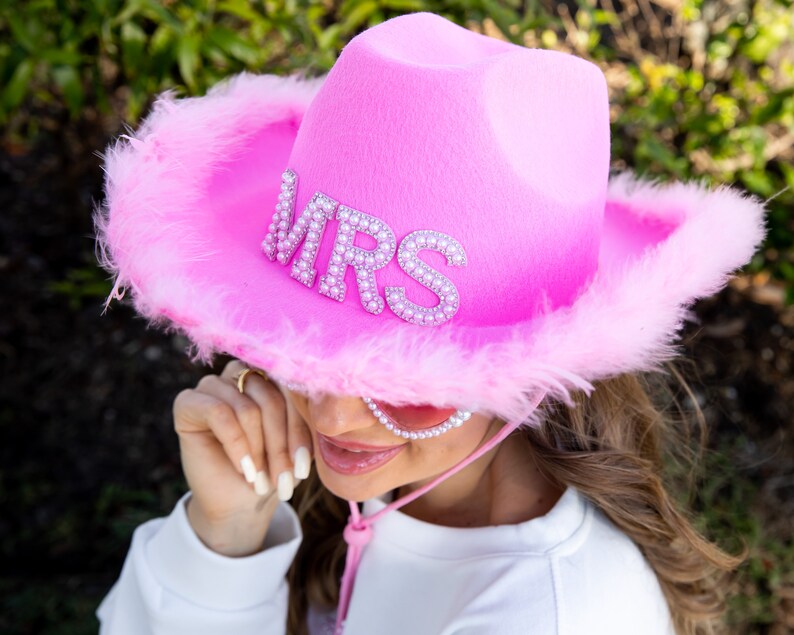May include: A bright pink cowboy hat with the word "MRS" spelled out in silver rhinestones. The hat has a fluffy, feathered trim and a pink chin strap. A fun accessory for a bachelorette party or wedding.