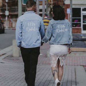 Puede incluir: Chaquetas vaqueras azul claro con las palabras "Mr. Dahlor" y "Mrs. Dahlor" en escritura blanca. Las chaquetas tienen detalles desgastados. La mujer lleva una falda de encaje blanco y el hombre pantalones negros. Caminan por una acera de ladrillos.