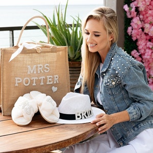 May include: A burlap tote bag with the text "MRS POTTER" in rhinestones. The bag is next to a pair of white fluffy slippers with rhinestone embellishments and a white straw hat with a black band and the word "BRIDE" in rhinestones.