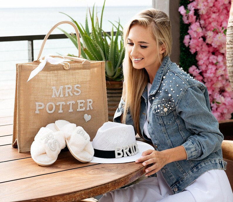 May include: A burlap tote bag with the text "MRS POTTER" in rhinestones. The bag is next to a pair of white fluffy slippers with rhinestone embellishments and a white straw hat with a black band and the word "BRIDE" in rhinestones.