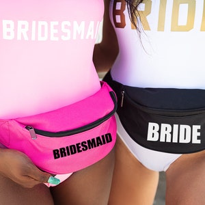 Può includere: Due donne indossano costumi da bagno bianchi a pezzo unico con marsupi neri. Il marsupio a sinistra dice "BRIDESMAID" in lettere nere e il marsupio a destra dice "BRIDE" in lettere bianche.