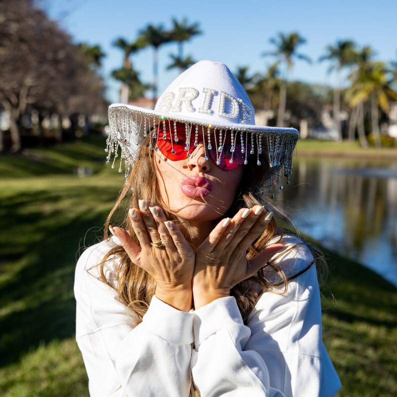 May include: A woman wearing a white hat with the word "BRIDE" on it and red heart-shaped sunglasses. She is blowing a kiss with both hands.