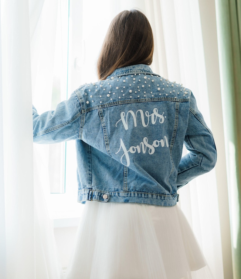 K&ouml;nnte beinhalten: Eine blaue Jeansjacke mit Perlenbesatz an den Schultern und dem Schriftzug "Mrs. Johnson" in Wei&szlig; auf dem R&uuml;cken.
