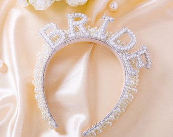May include: A white pearl headband with the word "BRIDE" spelled out in silver letters. The headband is on a light beige satin background.