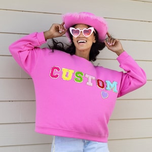 May include: A pink sweatshirt with the word "CUSTOM" in colorful letters. The sweatshirt has a relaxed fit and is worn by a person wearing a pink cowboy hat and heart-shaped sunglasses.