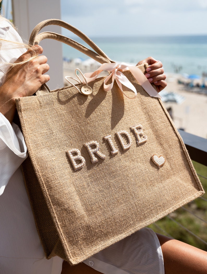 May include: A brown burlap tote bag with a pink ribbon and the word "BRIDE" in white pearl-like letters. A small heart is sewn on the bag.