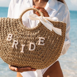 May include: A straw tote bag with a white ribbon and the word "BRIDE" spelled out in white letters with pearl accents.
