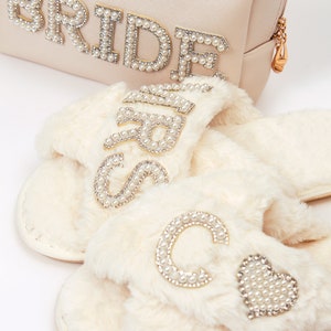 May include: A set of white faux fur slippers with pearl embellishments. One slipper has the word "Bride" spelled out in pearls, and the other slipper has a large "C" and a heart, both made of pearls.
