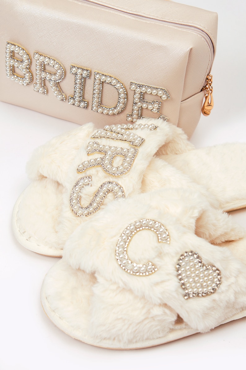 May include: A set of white faux fur slippers with pearl embellishments. One slipper has the word "Bride" spelled out in pearls, and the other slipper has a large "C" and a heart, both made of pearls.