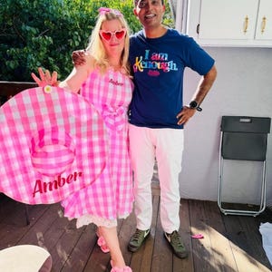 Peut inclure: Une femme porte une robe vichy rose et blanche et des lunettes de soleil en forme de c&oelig;ur, &agrave; c&ocirc;t&eacute; d'un homme en t-shirt bleu. La femme tient un grand anneau gonflable rose et blanc avec le nom "Amber". L'homme porte un pantalon blanc et des baskets vertes.