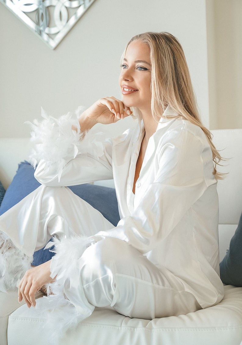 May include: A woman wearing a white satin pajama set with feather trim on the cuffs. She is sitting on a white couch with blue pillows.
