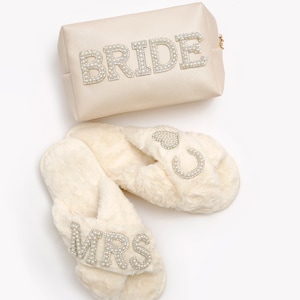 May include: A pair of white faux fur slippers with a criss-cross strap and pearl embellishments spelling "MRS" on one slipper and a heart and horseshoe on the other. A white satin cosmetic bag with pearl embellishments spelling "BRIDE" is also included.