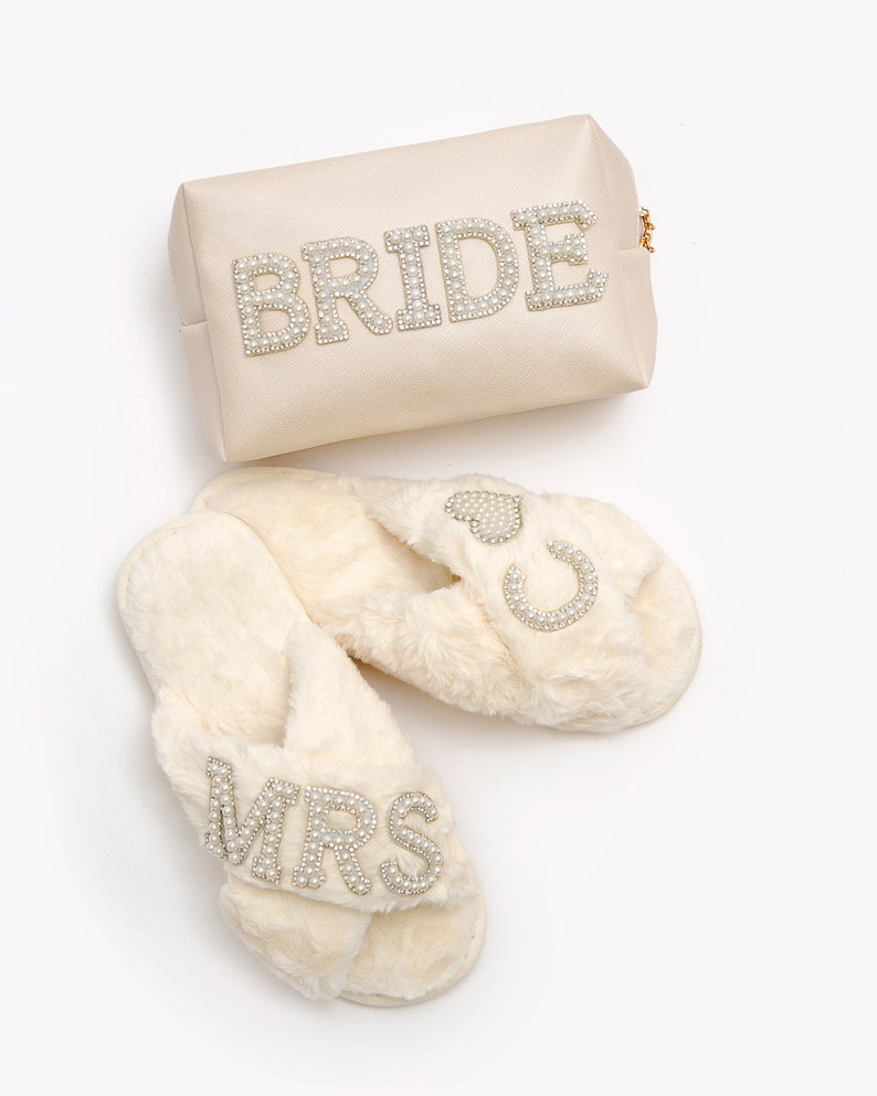 May include: A pair of white faux fur slippers with a criss-cross strap and pearl embellishments spelling "MRS" on one slipper and a heart and horseshoe on the other. A white satin cosmetic bag with pearl embellishments spelling "BRIDE" is also included.