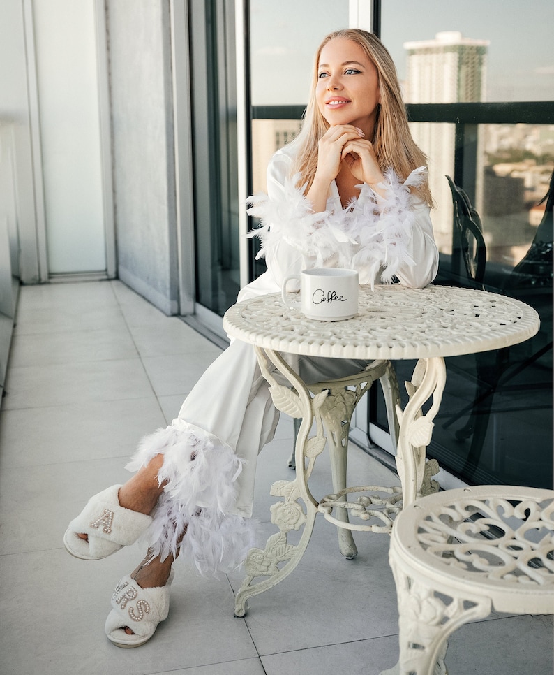 May include: A woman wearing a white satin robe with feather trim sits on a white patio chair with a white mug that says "Coffee". She is wearing white fluffy slippers with gold lettering that says "Boss".