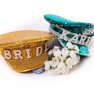 May include: Two sequined hats, one gold with the word "BRIDE" and one teal with the word "KARE", with a white flower bouquet.