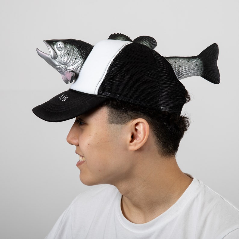 May include: A black and white baseball cap with a mesh back. A realistic-looking fish is attached to the front of the cap, with its head sticking out of the cap.