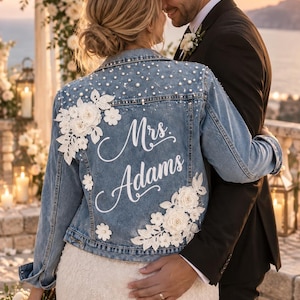 Può includere: Una giacca in denim blu con applicazioni in pizzo floreale bianco, perle bianche sparse e la scritta in corsivo bianco Mrs. Adams sul retro. La giacca presenta un colletto classico, maniche lunghe e una chiusura frontale con bottoni.