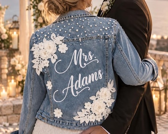May include: A blue denim jacket featuring white floral lace appliqués, scattered white pearls, and the white script text Mrs. Adams on the back. The jacket has a classic collar, long sleeves, and a button-front closure, worn over a white lace dress.