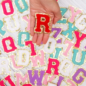 May include: A hand holding a red felt letter R with gold trim. The letter is surrounded by a variety of colorful felt letters with gold trim.