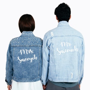 May include: A couple's denim jackets with personalized embroidery. The woman's jacket has pearl embellishments and says "Mrs. Saivouch" with the date "05.03.2023". The man's jacket says "Mr. Saivouch" with the date "05.03.2023".