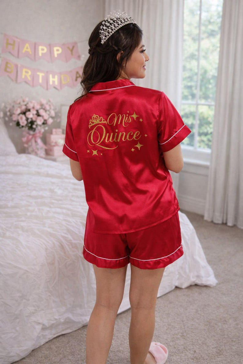 Puede incluir: Conjunto de pijama de sat&eacute;n rojo con ribete blanco. La parte superior presenta el texto dorado "Mis Quince" con un dise&ntilde;o de corona. Los pantalones cortos y la parte superior tienen ribete blanco. La imagen muestra a una persona usando el conjunto.