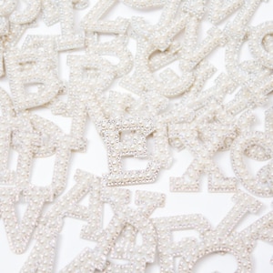 May include: A close-up of a collection of pearl-embellished letter patches. The letter "E" is in the center of the image, surrounded by other letters.