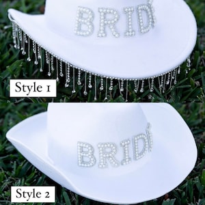May include: Two white cowboy hats with the word "BRIDE" spelled out in pearl beads. The top hat has a fringe of silver beads hanging from the brim. The bottom hat does not have a fringe.