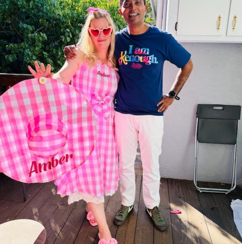 Puede incluir: Una mujer con un vestido de cuadros rosas y blancos y gafas de sol en forma de coraz&oacute;n est&aacute; de pie junto a un hombre con una camiseta azul. La mujer sostiene un gran flotador inflable rosa y blanco con el nombre "Amber". El hombre lleva un reloj y pantalones blancos.