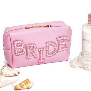 May include: Pink cosmetic bag with a zipper closure and the word "BRIDE" spelled out in pearl-embellished letters.