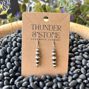 May include: Silver beaded dangle earrings displayed on a brown card with the text "THUNDER & STONE HANDMADE JEWELRY". The earrings are set against a backdrop of black beans.