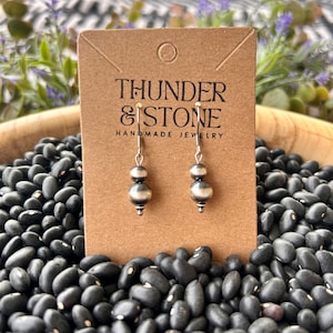 May include: Silver earrings with two round beads on each earring. The earrings are displayed on a brown card with the text "THUNDER & STONE HANDMADE JEWELRY".
