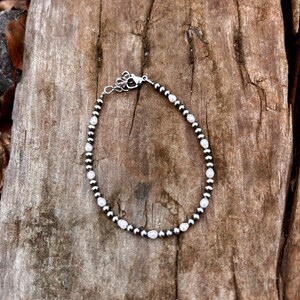 May include: A delicate bracelet featuring small, round silver beads interspersed with oval, white beads. The bracelet is secured with a silver clasp and rests on a textured, brown wooden surface. This piece of jewelry is suitable for everyday wear.