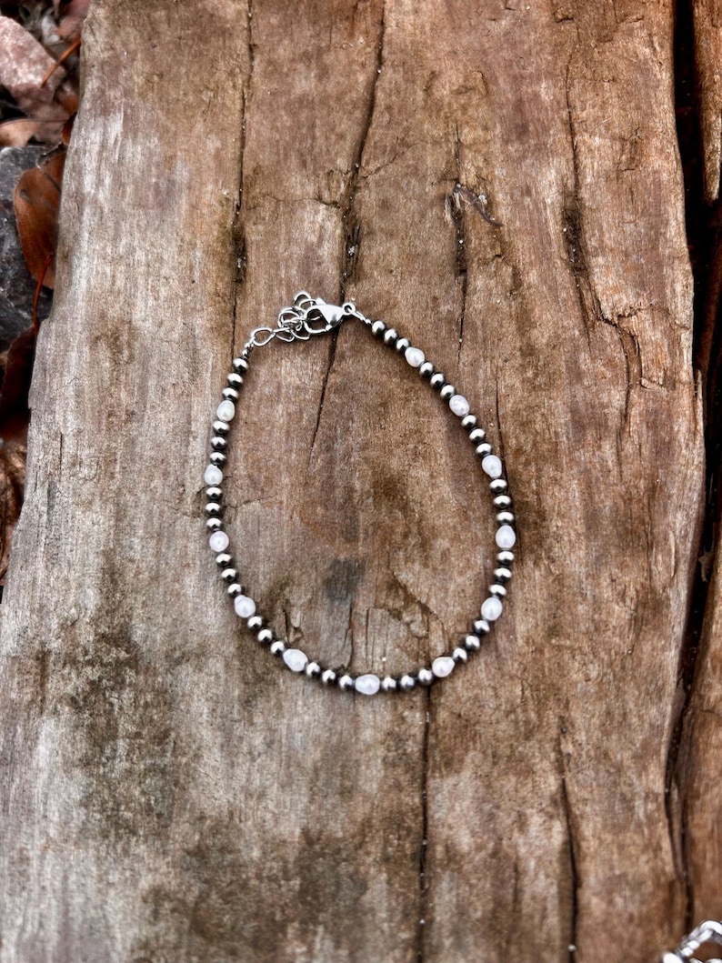 May include: A delicate bracelet featuring small, round silver beads interspersed with oval, white beads. The bracelet is secured with a silver clasp and rests on a textured, brown wooden surface. This piece of jewelry is suitable for everyday wear.