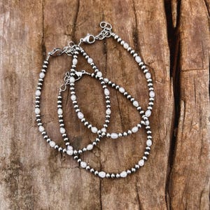 May include: Three silver bracelets with a mix of small, round, silver beads and white, oval-shaped beads. The bracelets are laid out on a textured, brown wooden surface. Each bracelet has a clasp closure.