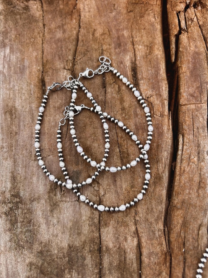 May include: Three silver bracelets with a mix of small, round, silver beads and white, oval-shaped beads. The bracelets are laid out on a textured, brown wooden surface. Each bracelet has a clasp closure.
