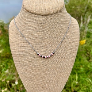 May include: A delicate necklace with a silver chain and a row of small pink and silver beads. The necklace is displayed on a burlap-covered bust, with a blurred natural background of water and greenery.