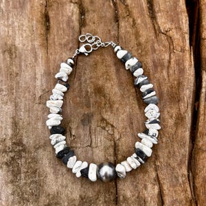 May include: A beaded bracelet featuring a mix of white, black, and gray stones. The bracelet has a silver-colored clasp and a large, round, silver-colored bead in the center. The stones are irregularly shaped and arranged in a pattern.